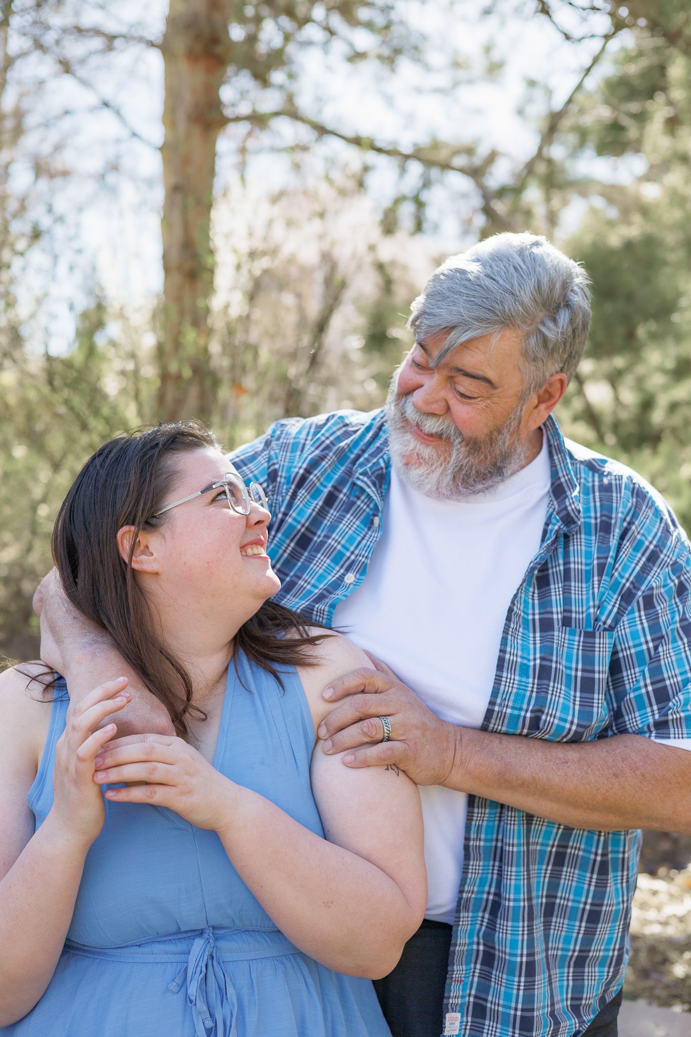 Older bearded man with gray hair embraces young woman with glasses outdoors in a wooded park setting.
