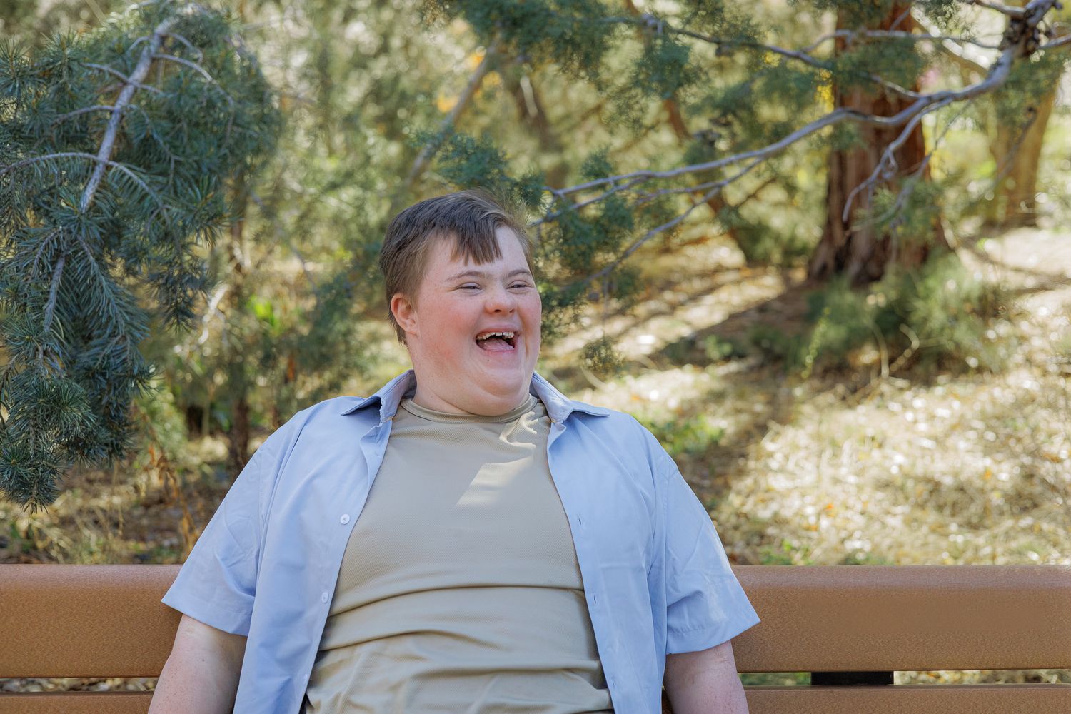 A cheerful young man with Down syndrome laughs while sitting on a bench outdoors.