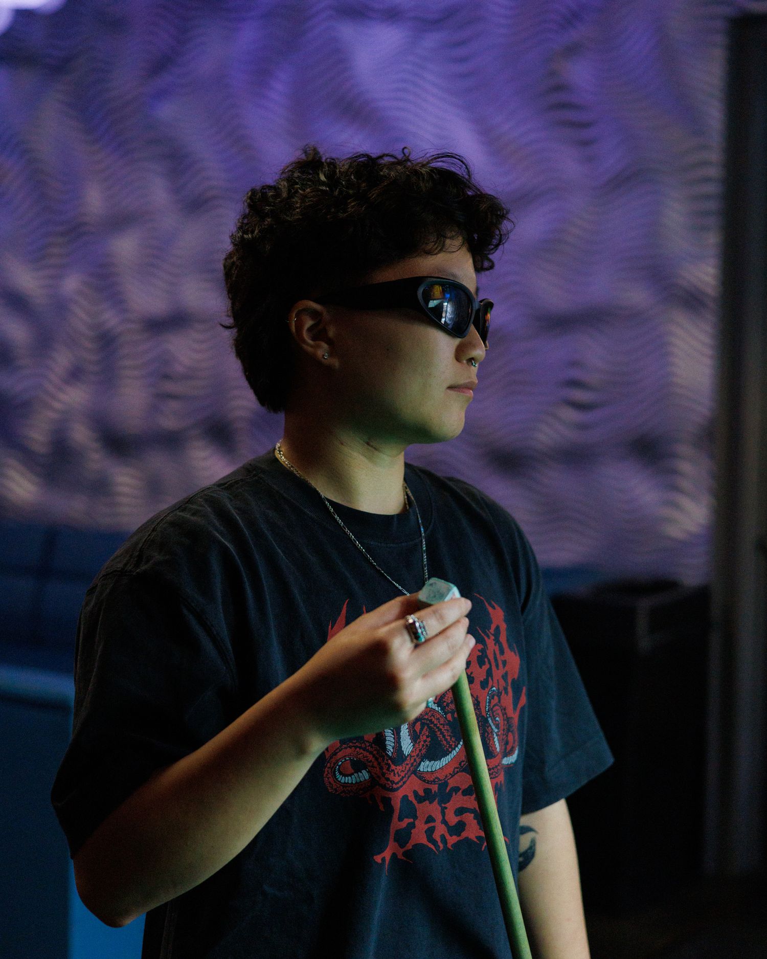 Young person with curly hair wearing futuristic sunglasses holding microphone under purple stage lighting.