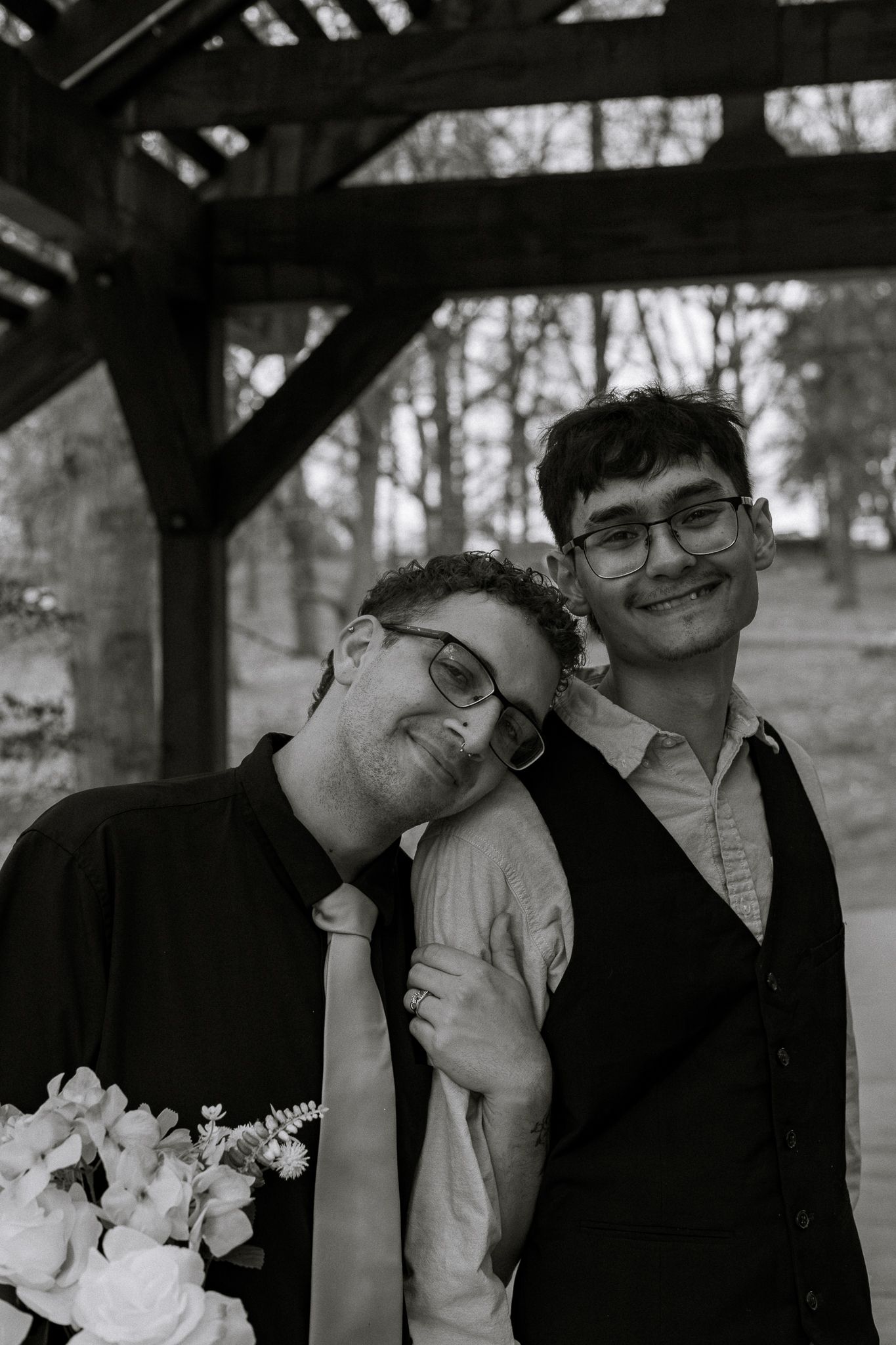 Black and white photo of two smiling men in formal wear holding flowers under a wooden structure.