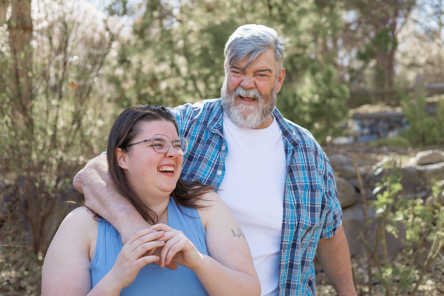 An older bearded man with gray hair embraces a younger woman with glasses outdoors among trees.