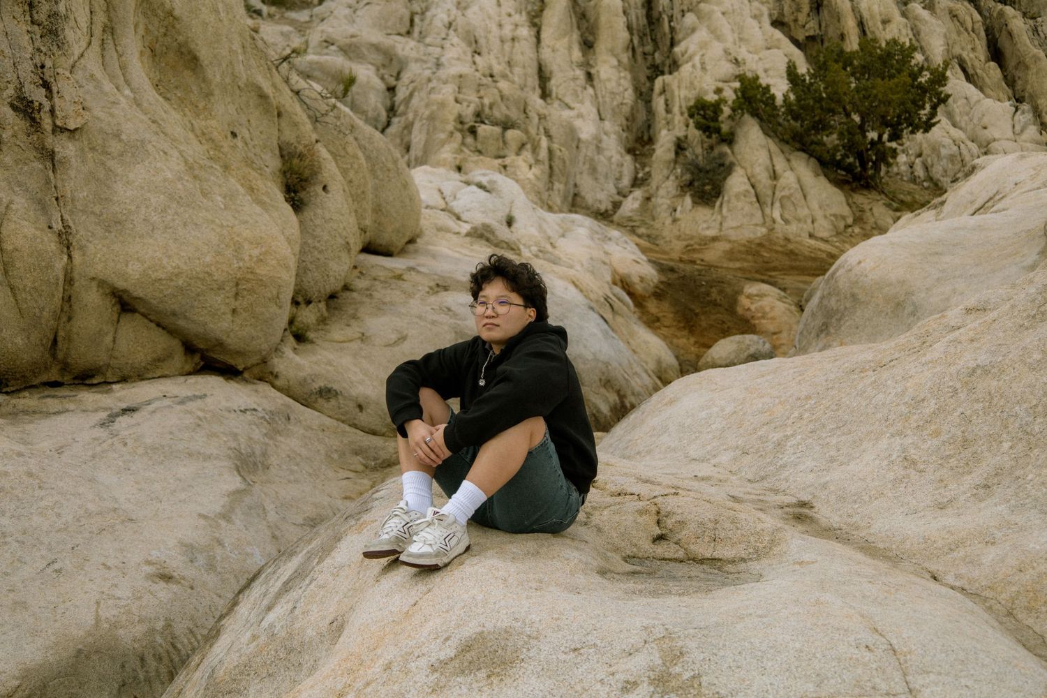 A young person with glasses sits relaxed on large rocky boulders in a desert landscape.