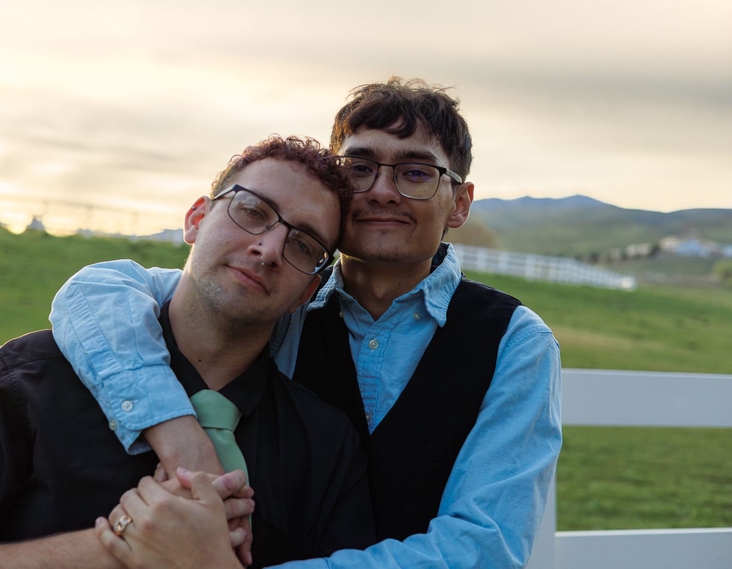 Two men with glasses embrace tenderly at sunset near a white fence with mountain views.