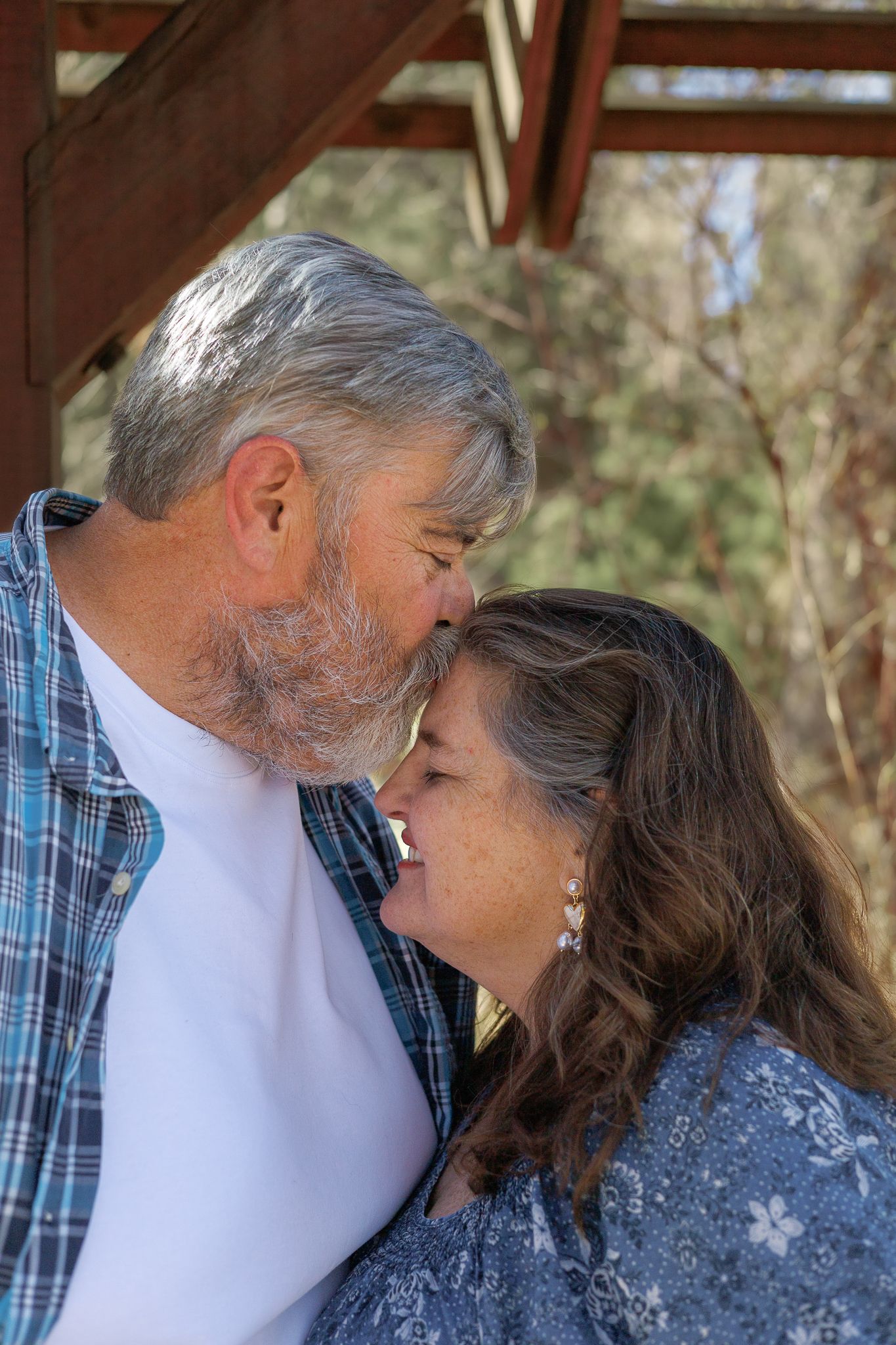 Older couple sharing a tender nose-to-nose moment outdoors under a wooden structure.