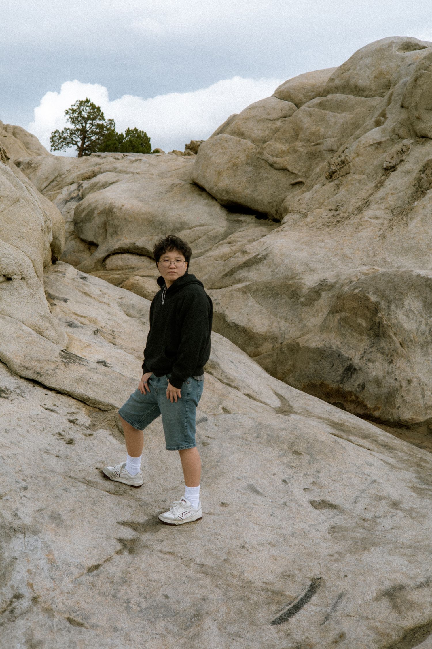 Young person in black hoodie and shorts posing on rocky terrain with cloudy sky background.