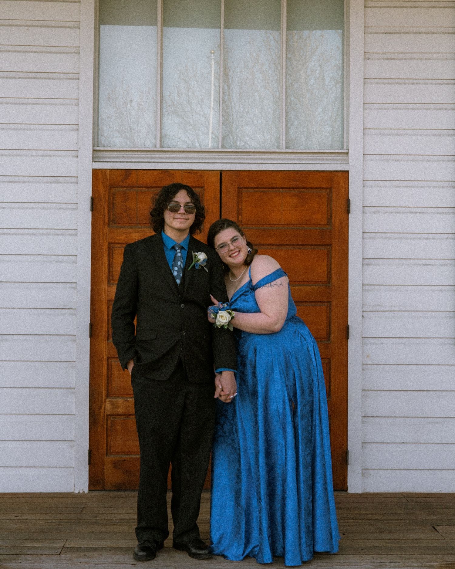 A couple dressed formally poses in front of wooden doors, the woman wearing a blue satin gown.