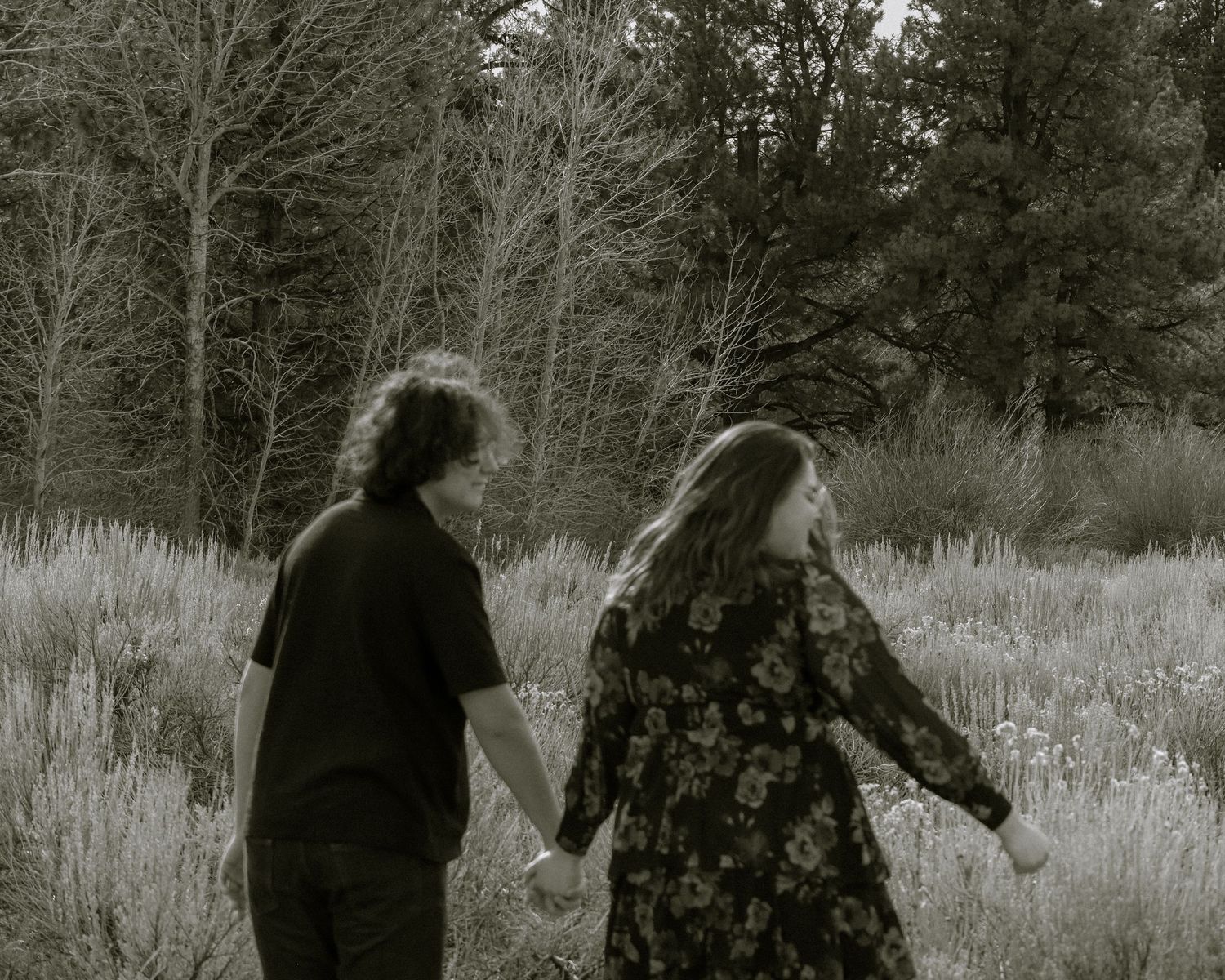Black and white photo of young couple holding hands walking through a meadow with tall trees.