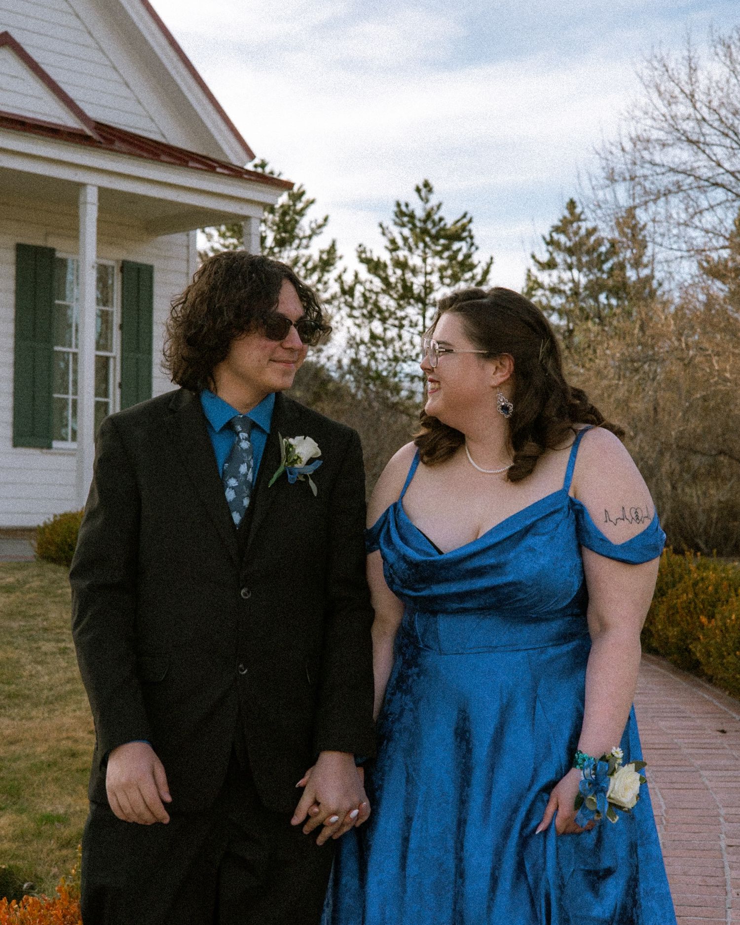 A couple in formal attire holds hands and smiles at each other outside a white building.