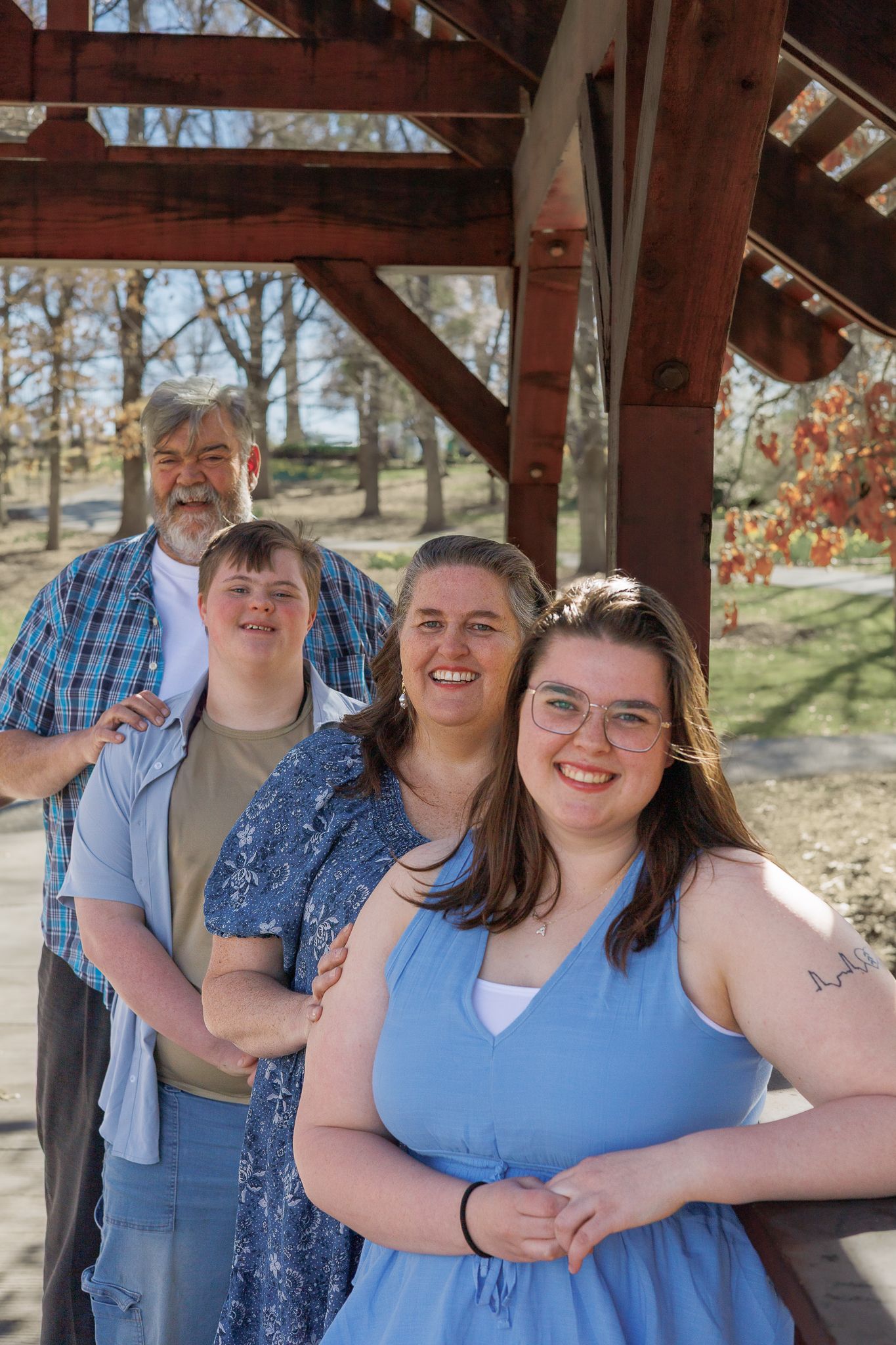 Family of four smiling together under a rustic wooden covered bridge in an autumn park setting.