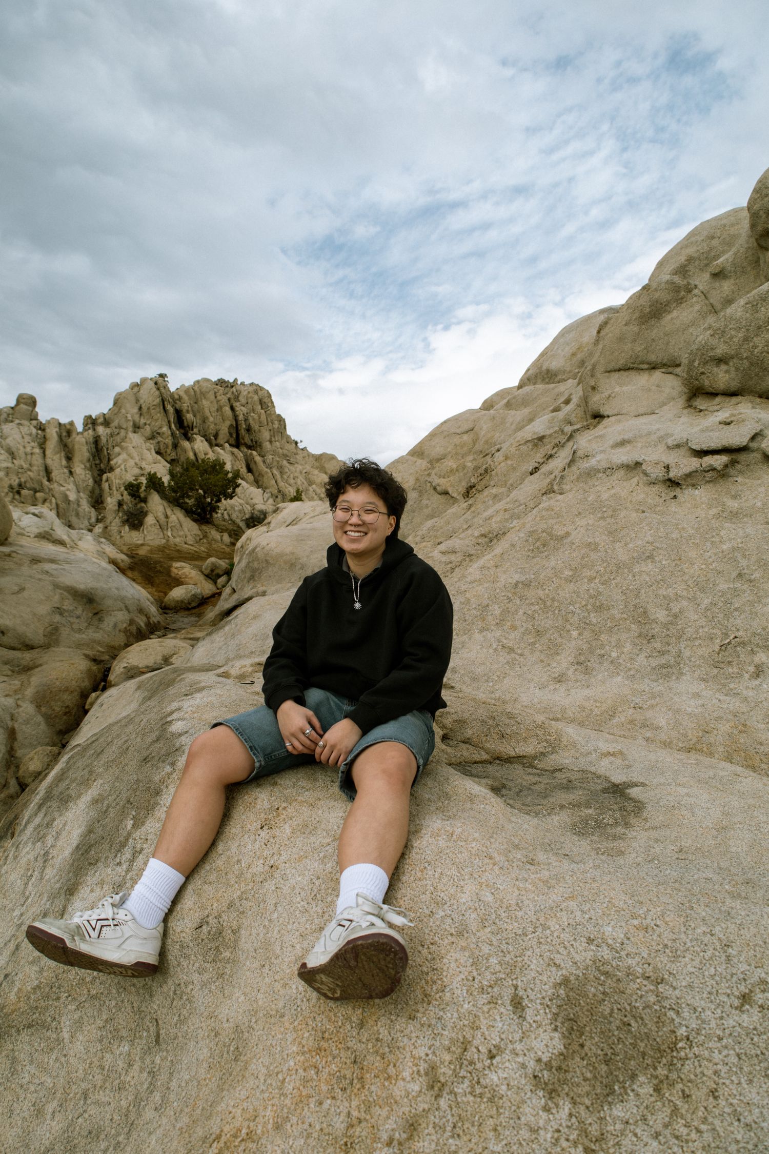 Smiling young person in black hoodie and shorts sitting on large rock with rocky landscape behind.