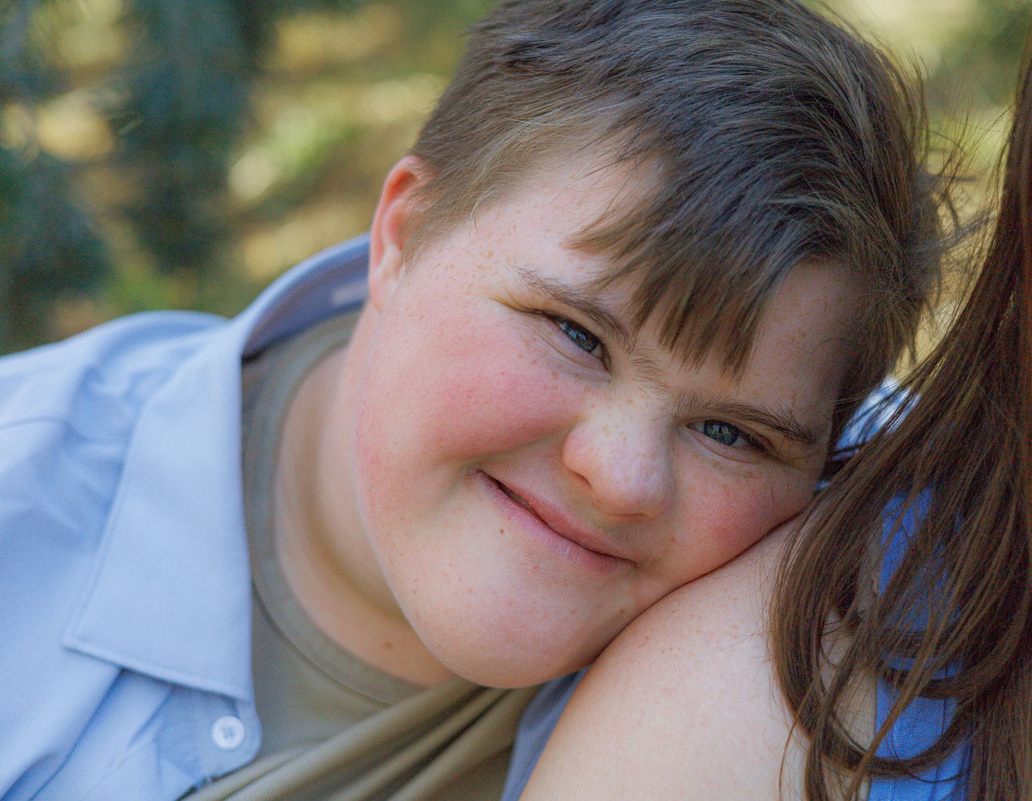 Smiling young boy with Down syndrome rests his head on someone's shoulder in an outdoor setting.