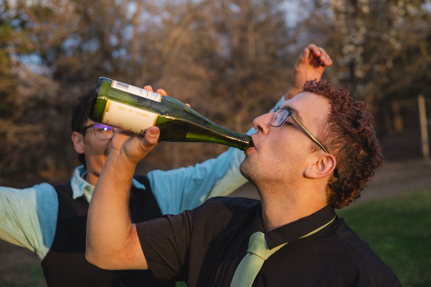 Man in glasses drinks champagne from a bottle outdoors while another person stands behind him.