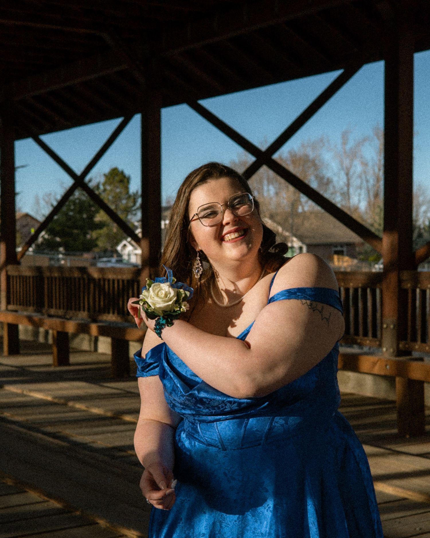 Young woman in blue off-shoulder prom dress with wrist corsage posing on wooden bridge outdoors.