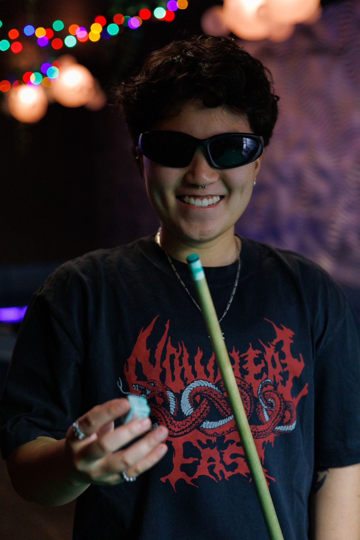 Smiling young person in sunglasses and black graphic tee holds a pool cue under colorful string lights.