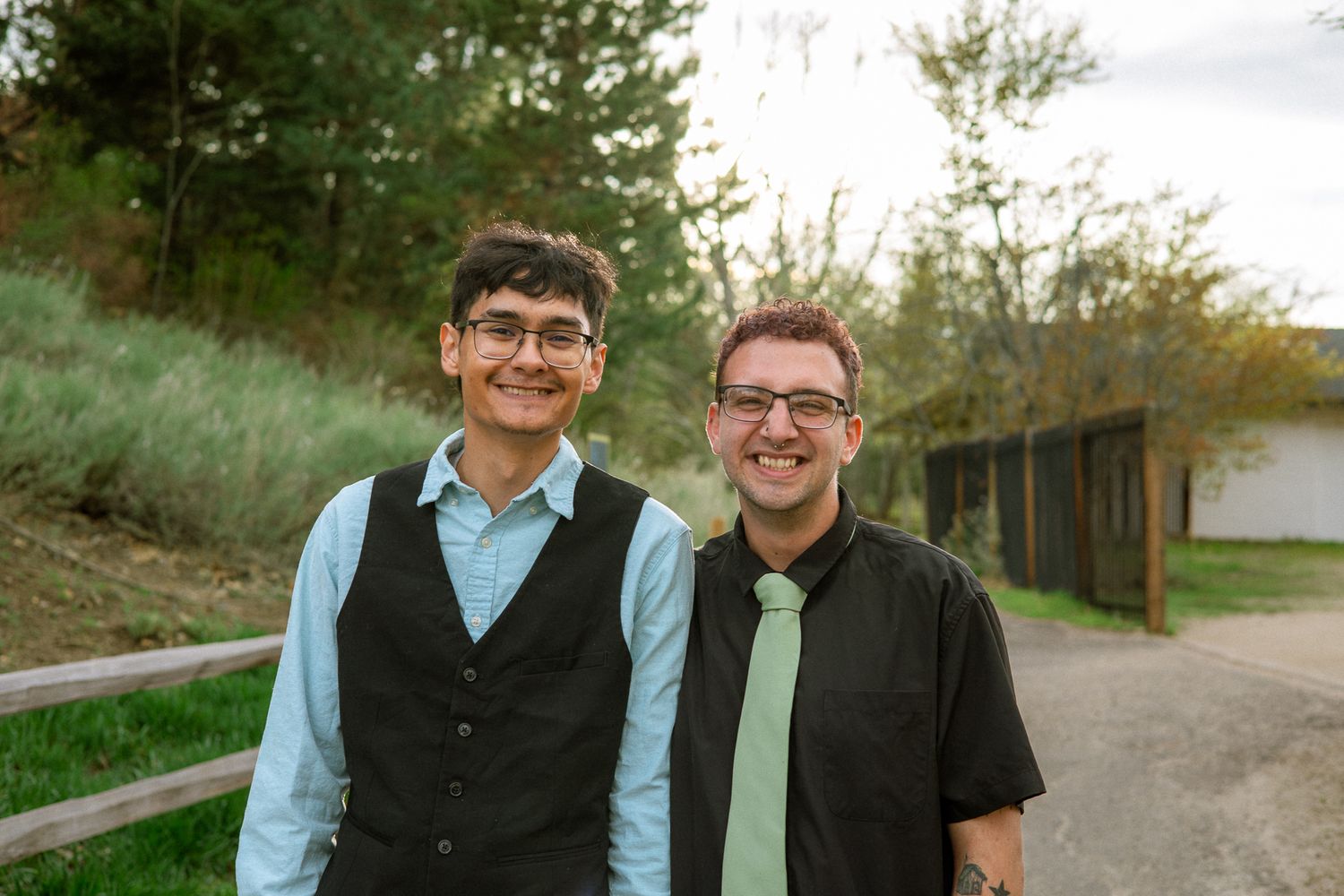 Two smiling men in formal attire stand together on a garden path surrounded by autumn trees.