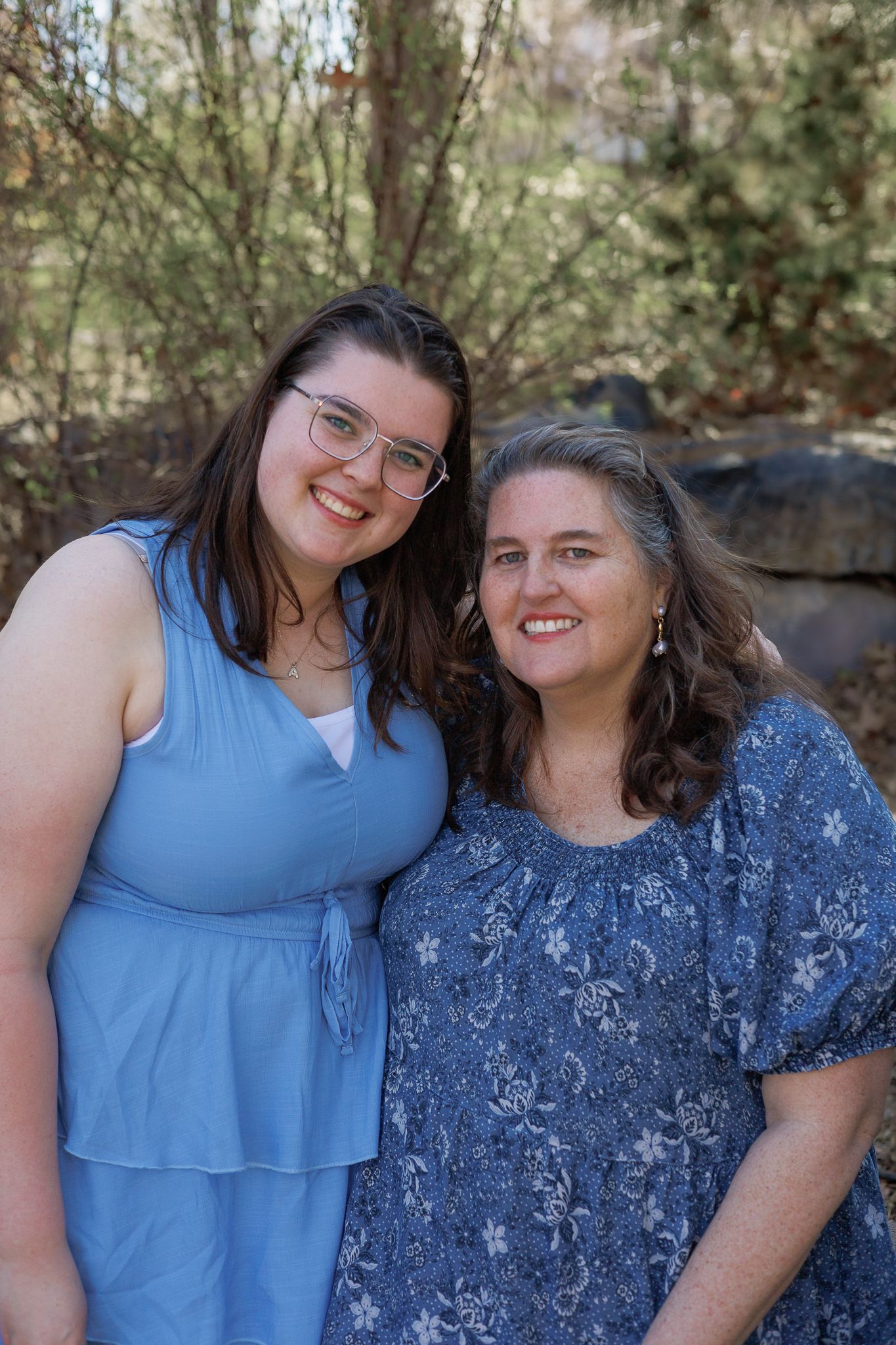 Two women in blue outfits smiling together outdoors with trees and rocks in the background.