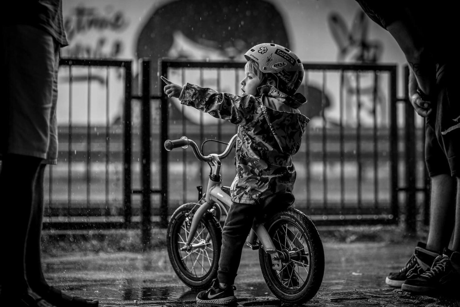 Black and white photo of a young child riding a balance bike in the rain wearing a helmet.