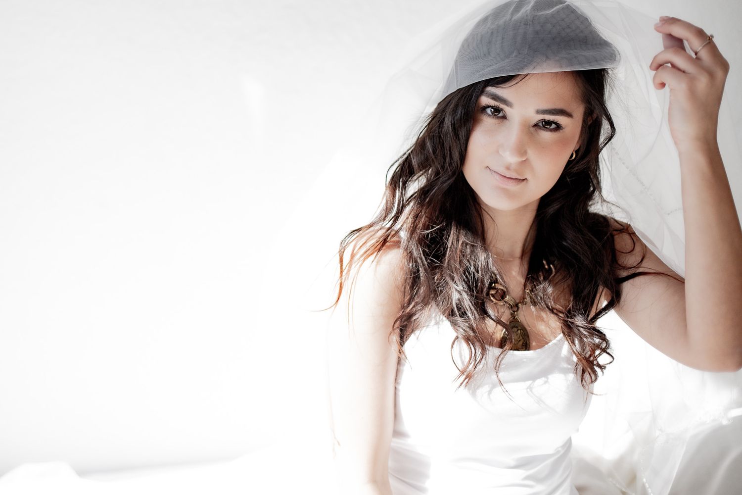 A beautiful bride with dark curly hair lifts her white veil, wearing a white dress in a bright studio setting.