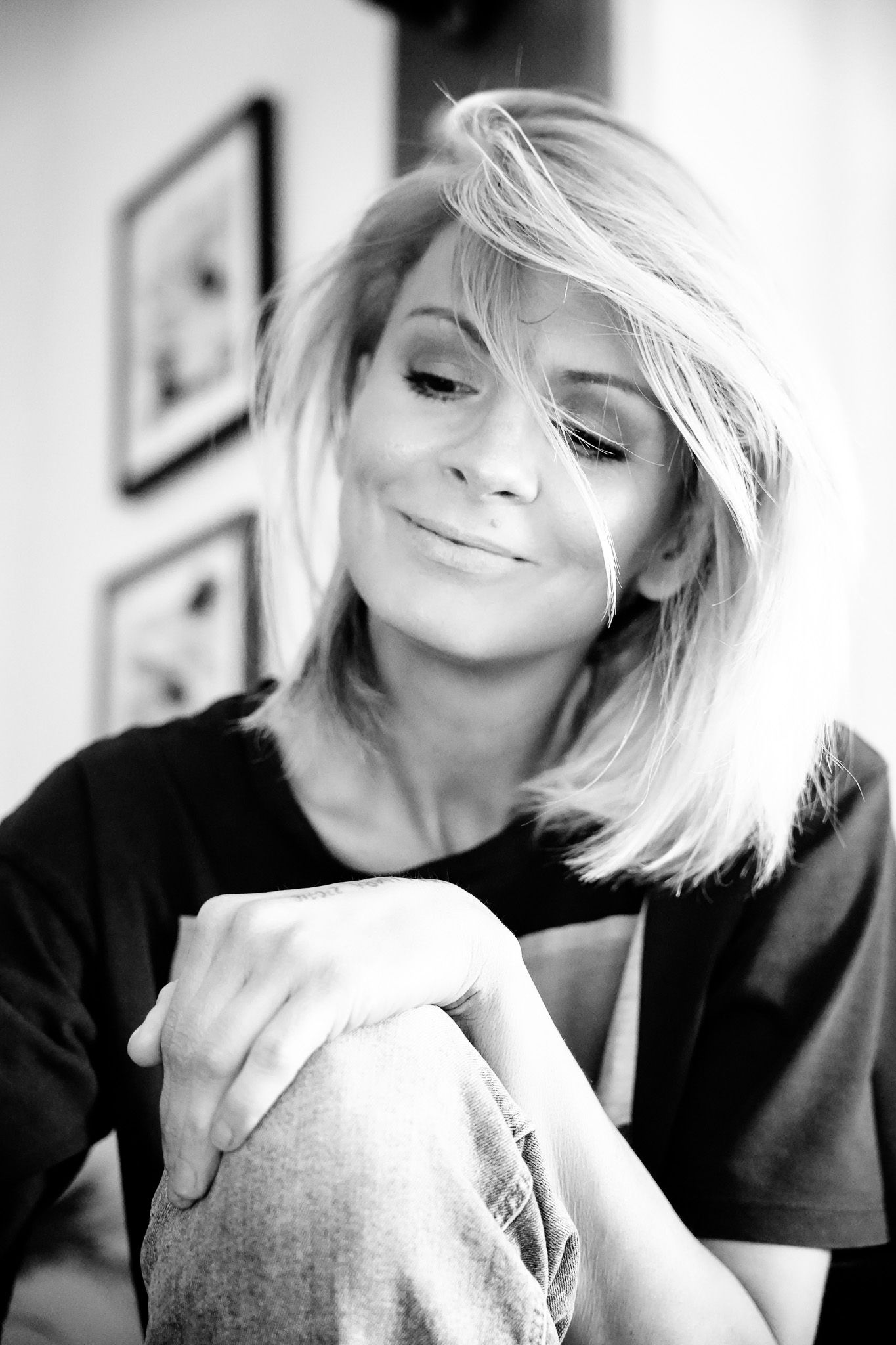 Black and white portrait of a smiling blonde woman with short hair, wearing a dark top, sitting with knees up.
