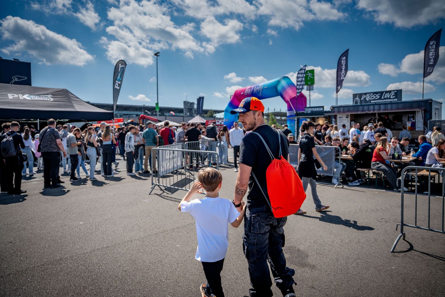 An adult and child walk hand-in-hand toward a busy outdoor car show venue on a sunny day.