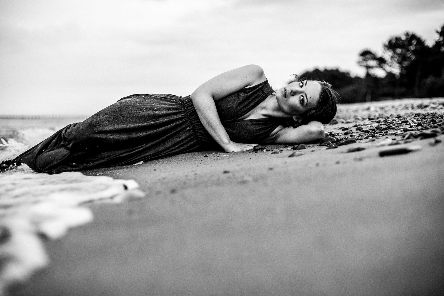 Black and white photo of a woman in a dark dress lying on a beach near the water.