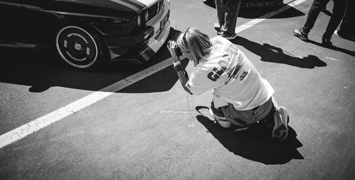 Black and white photo of a person crouching to photograph a lowered classic car at an outdoor car show.