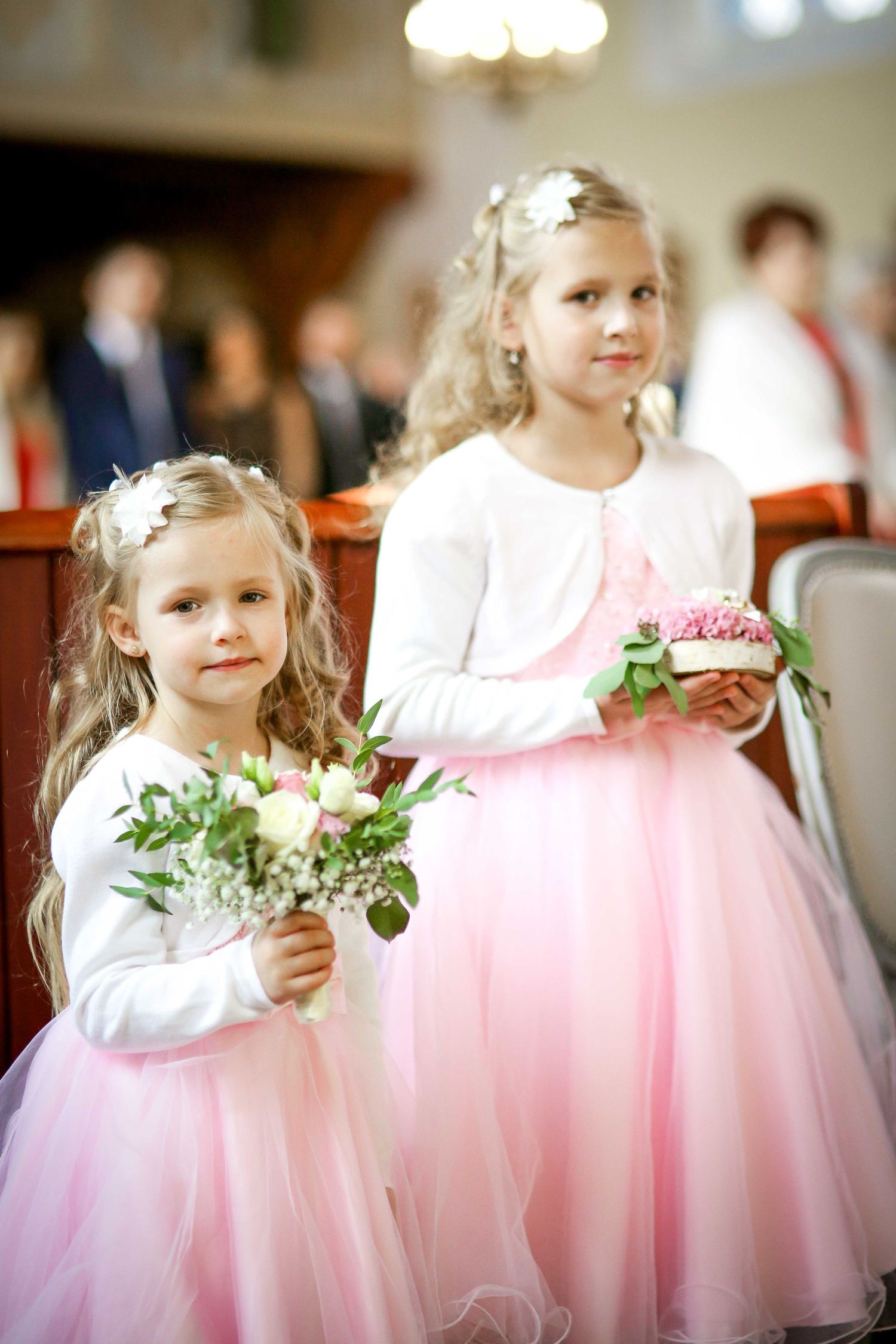 Two young flower girls in pink tulle dresses holding bouquets during a church wedding ceremony.