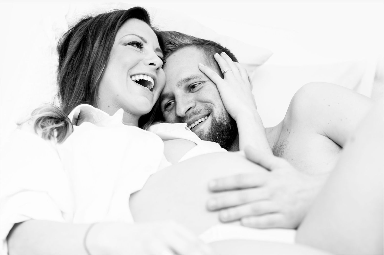 Black and white photo of laughing pregnant couple sharing a tender moment together in bed.