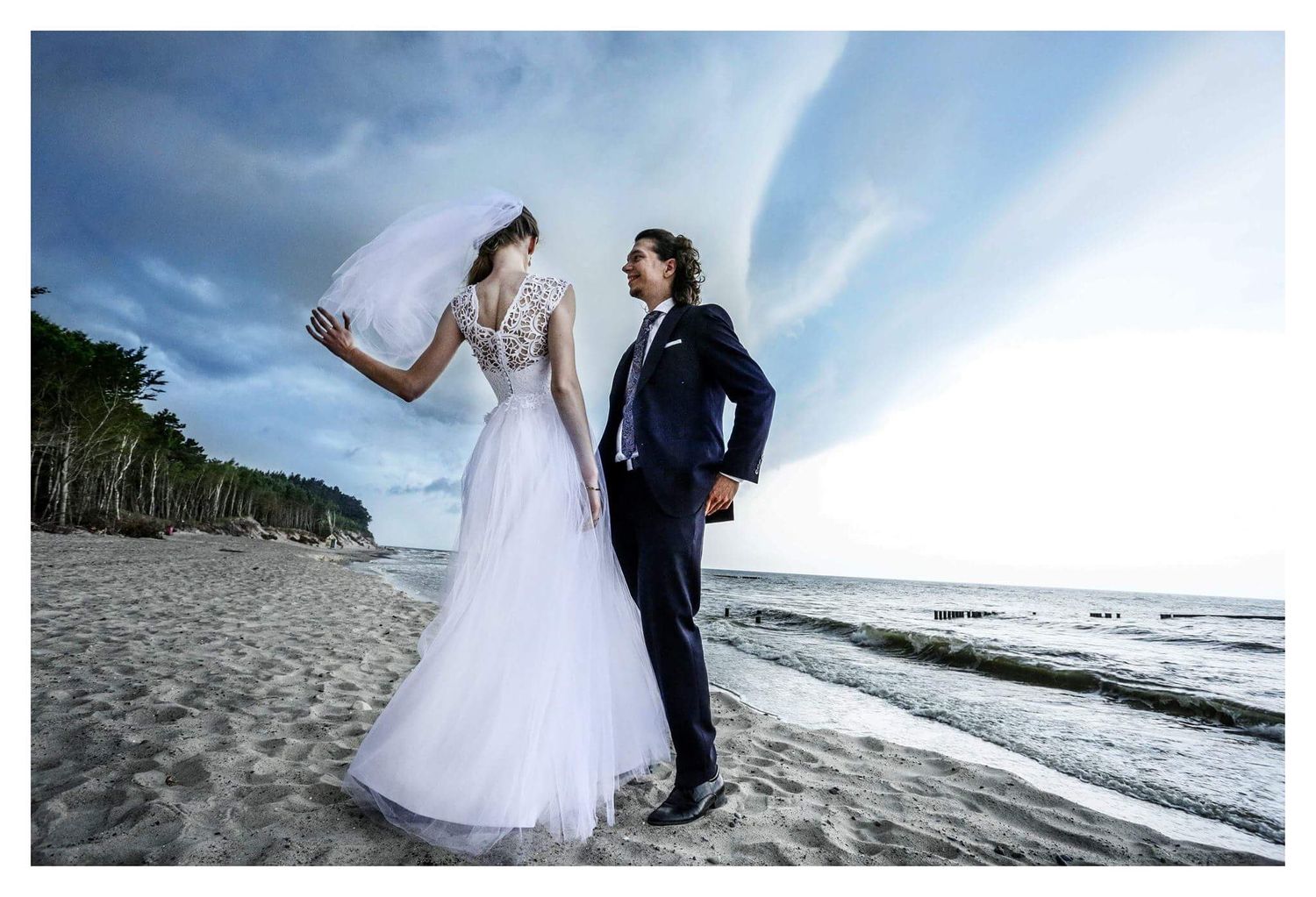 Bride and groom standing on a sandy beach with dramatic cloudy sky and trees in background.
