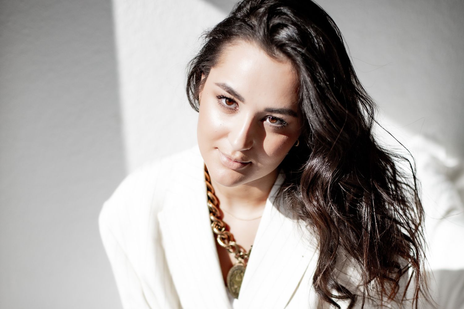 Young brunette woman in white blazer with gold chain necklace posing in soft natural light.