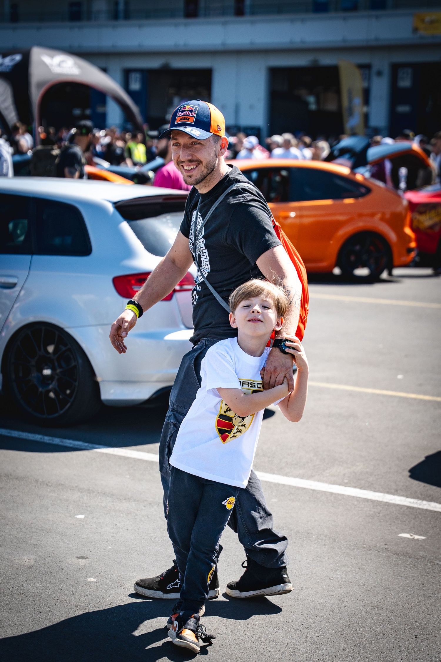 A smiling man in a Red Bull cap playfully carries a young boy at a sunny outdoor automotive event.