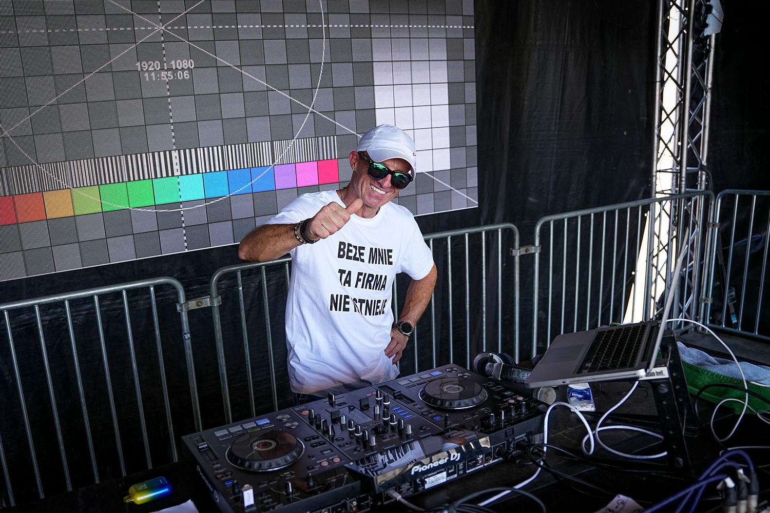 DJ performs at outdoor festival behind turntables wearing white t-shirt and cap in daylight.