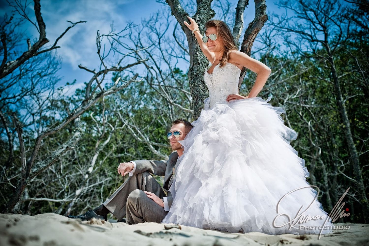 Bride in layered white gown stands over groom on sandy beach surrounded by bare trees.