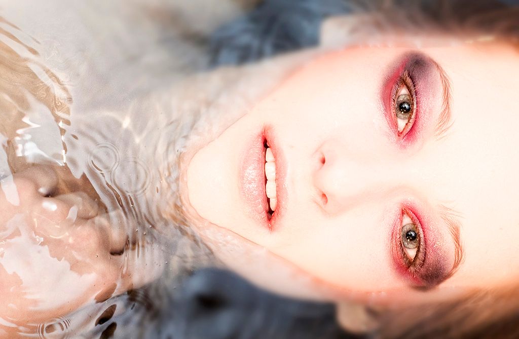 Close-up portrait of a woman with hazel eyes and rosy makeup lying in shallow water.