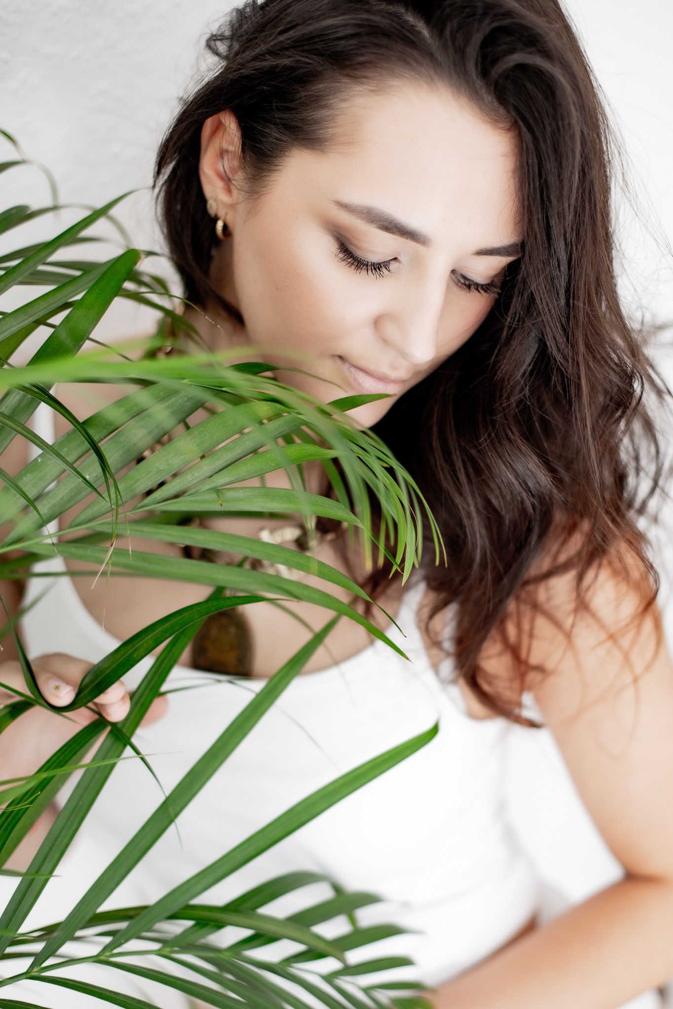 Brunette woman with long lashes looking down beside lush green tropical palm leaves indoors.