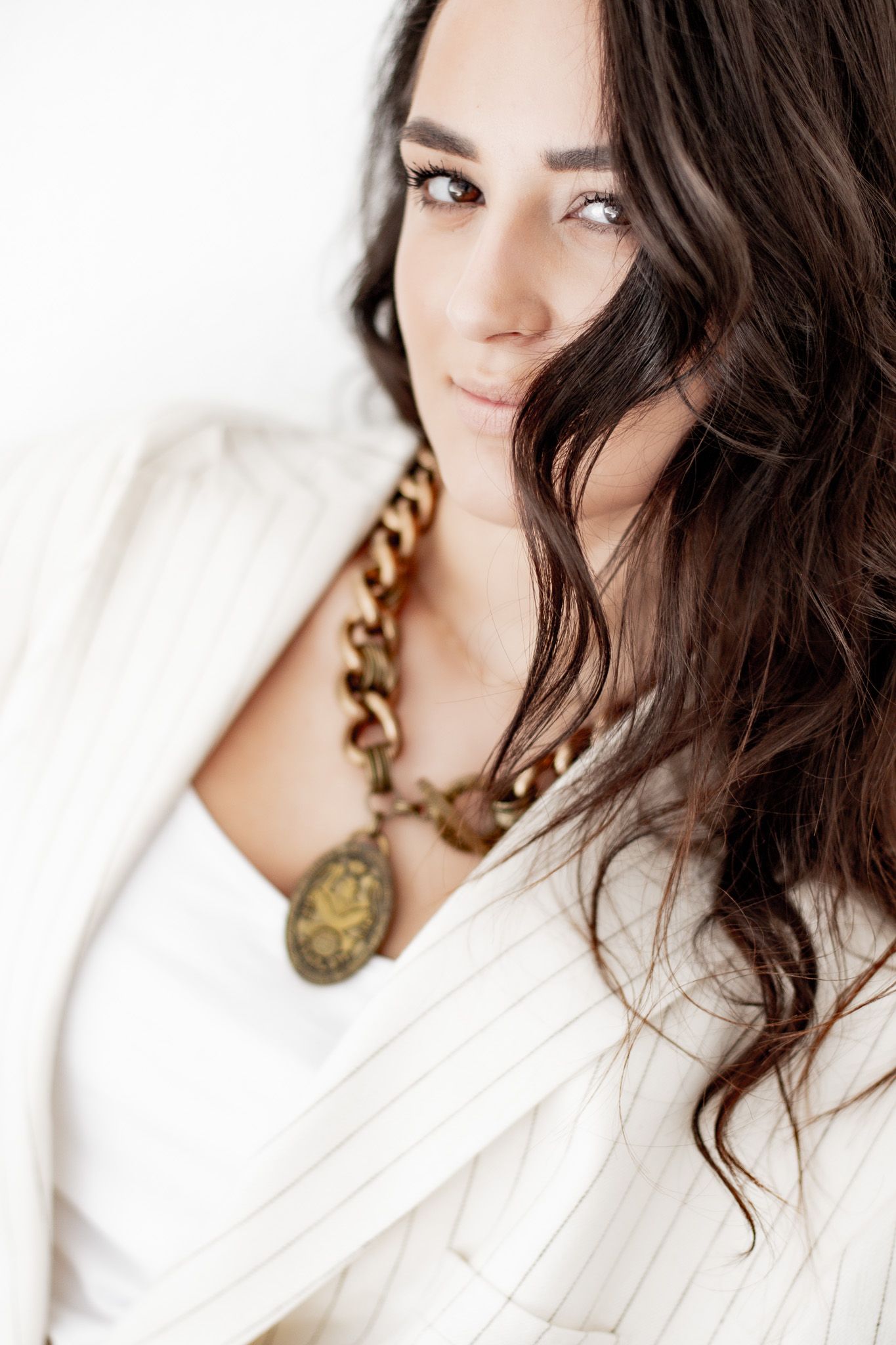 A brunette woman with wavy hair wearing a white pinstripe blazer and gold chain necklace poses in soft light.