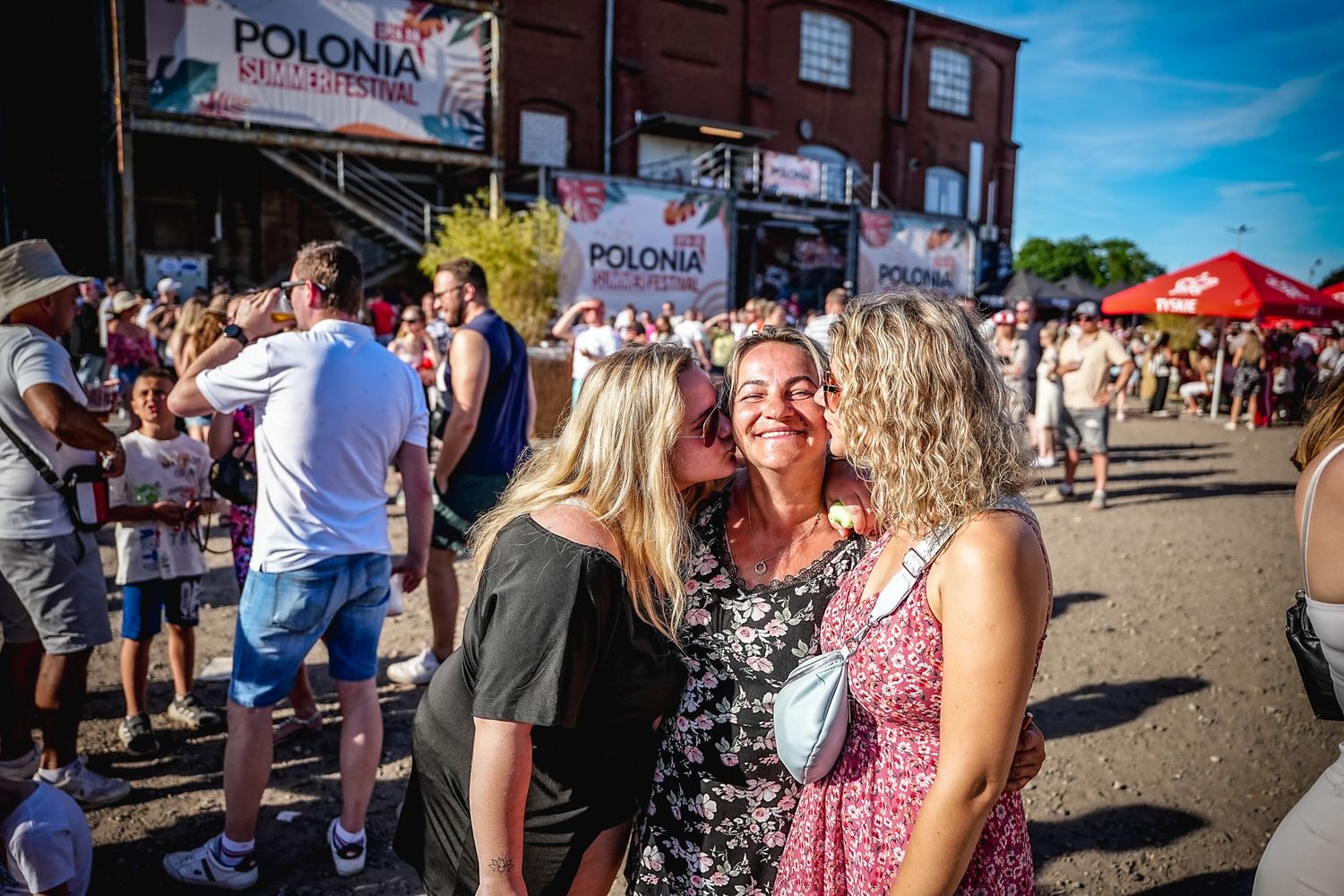 Three women share kisses at Polonia Summer Festival outdoor event with crowd in background.