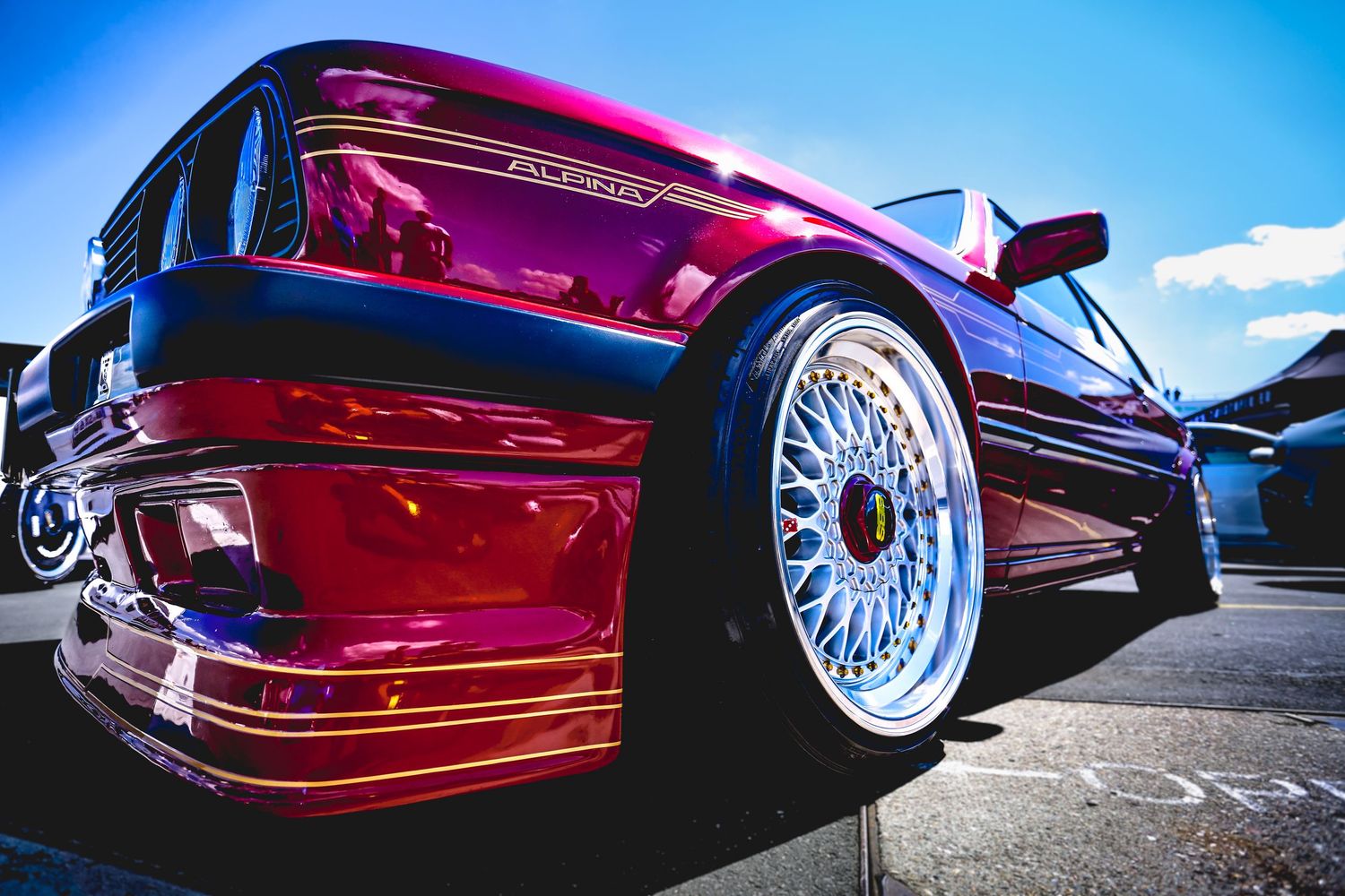Lowered burgundy BMW E30 with BBS wheels and wide body kit at a car show.
