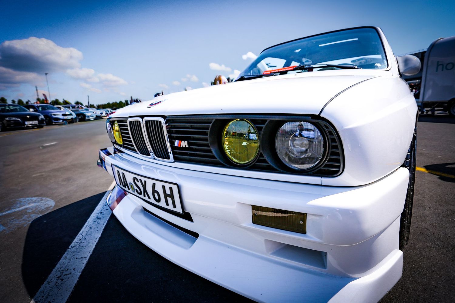 White BMW M3 E30 with yellow headlights and M badge on a parking lot under blue sky.