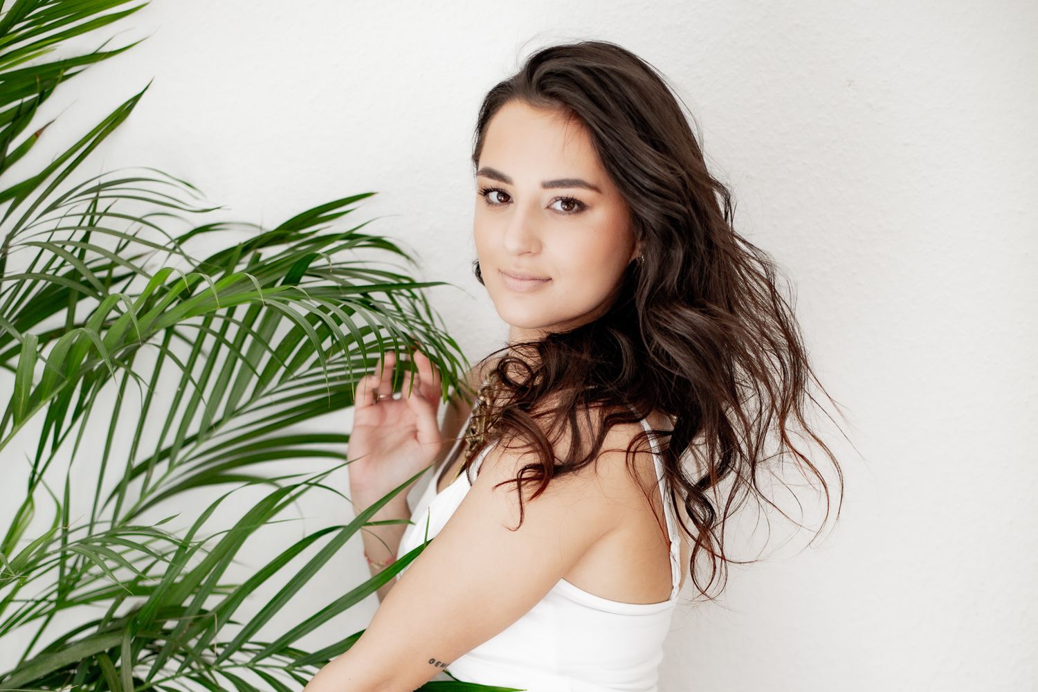 A young brunette woman with wavy hair poses beside tropical palm leaves against a white wall in a white top.