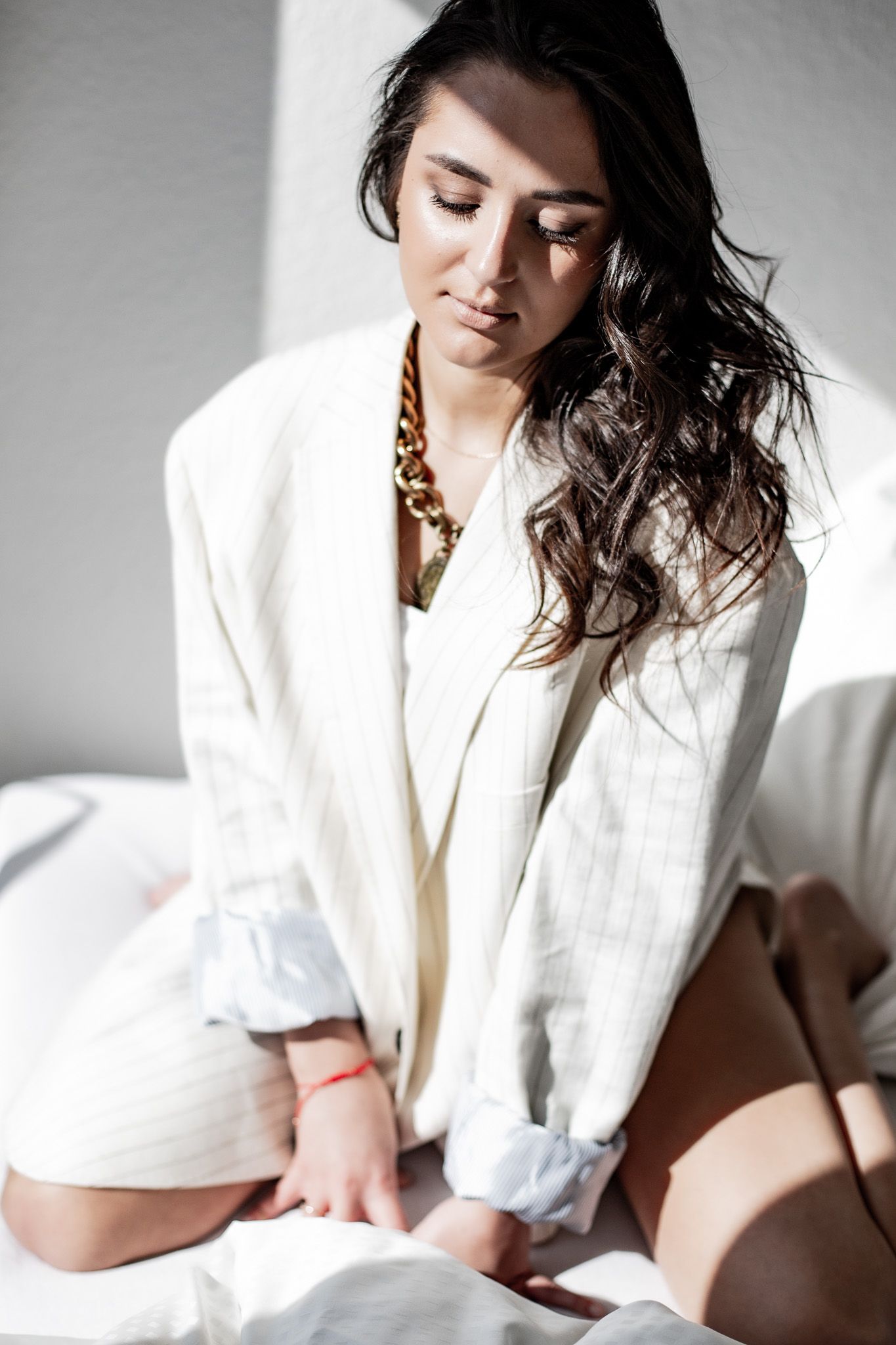 Brunette woman in white blazer and shorts sitting on bed in sunlit room with gold necklace.