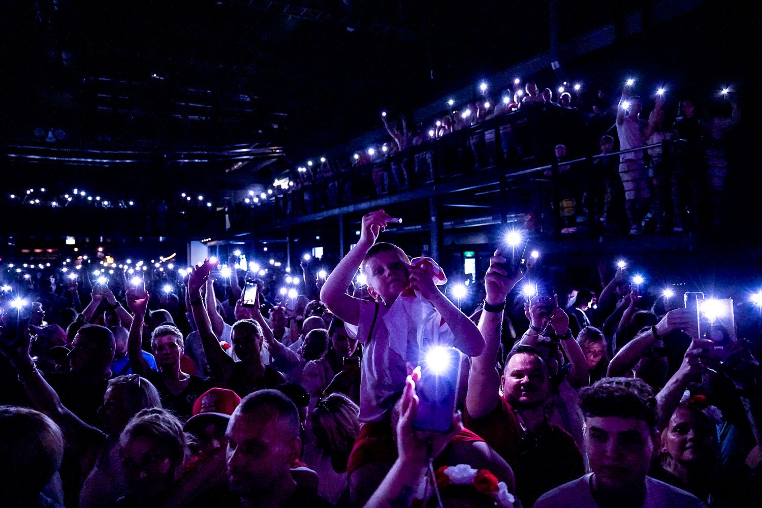 Crowd of concert fans holding up glowing phone lights in a packed dark arena during a live music event.Polonia music festival