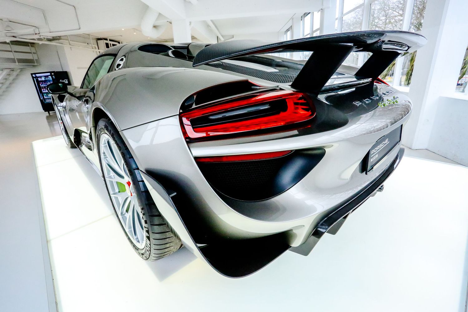 Rear quarter view of a silver Porsche 918 Spyder with large wing in a white showroom.