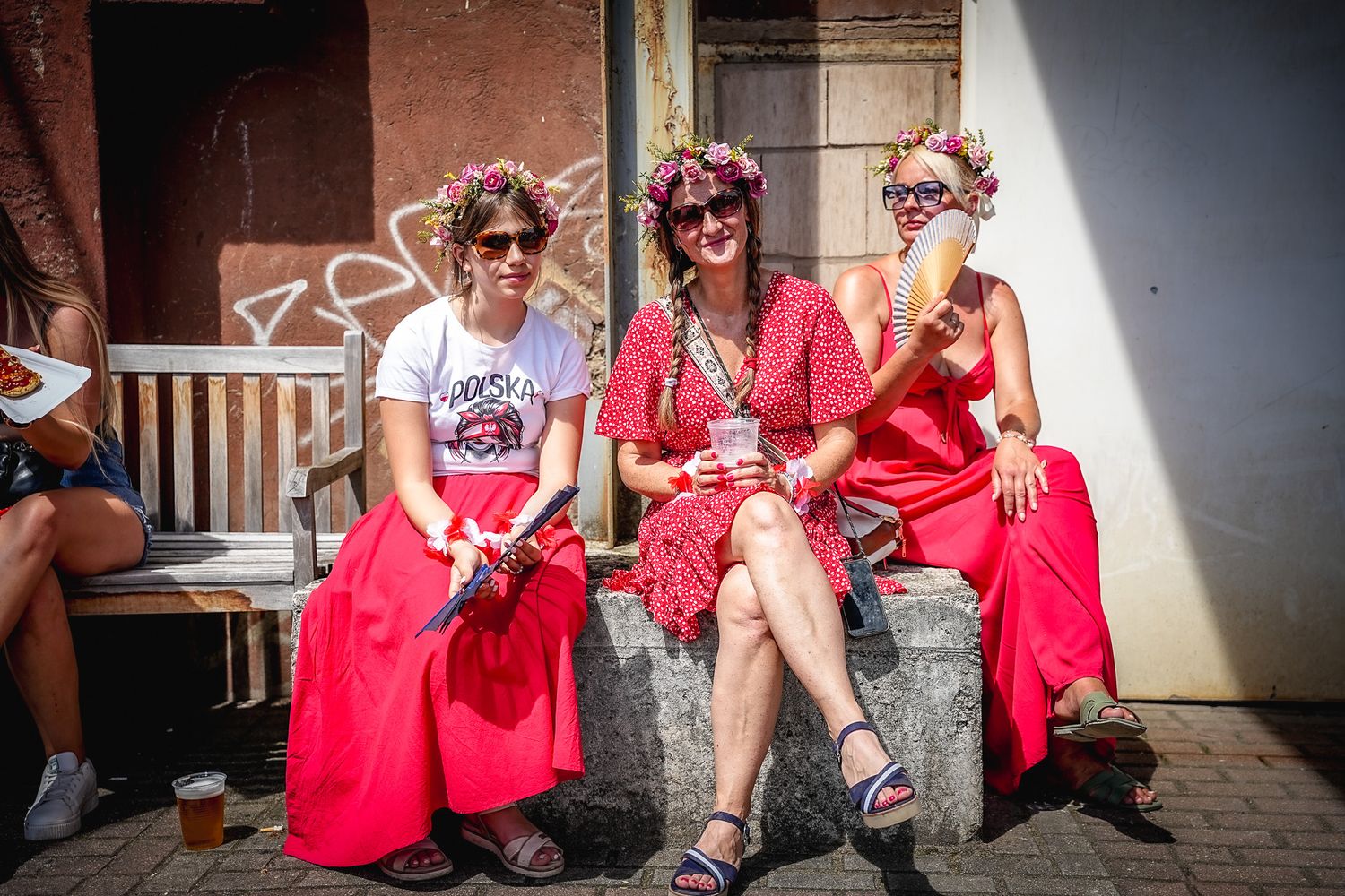 Three women in red outfits with flower crowns sit together at sunny outdoor summer festival.