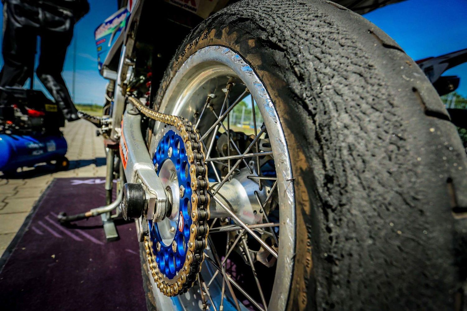 Close-up of motorcycle rear wheel with chain and blue hub at racing event.