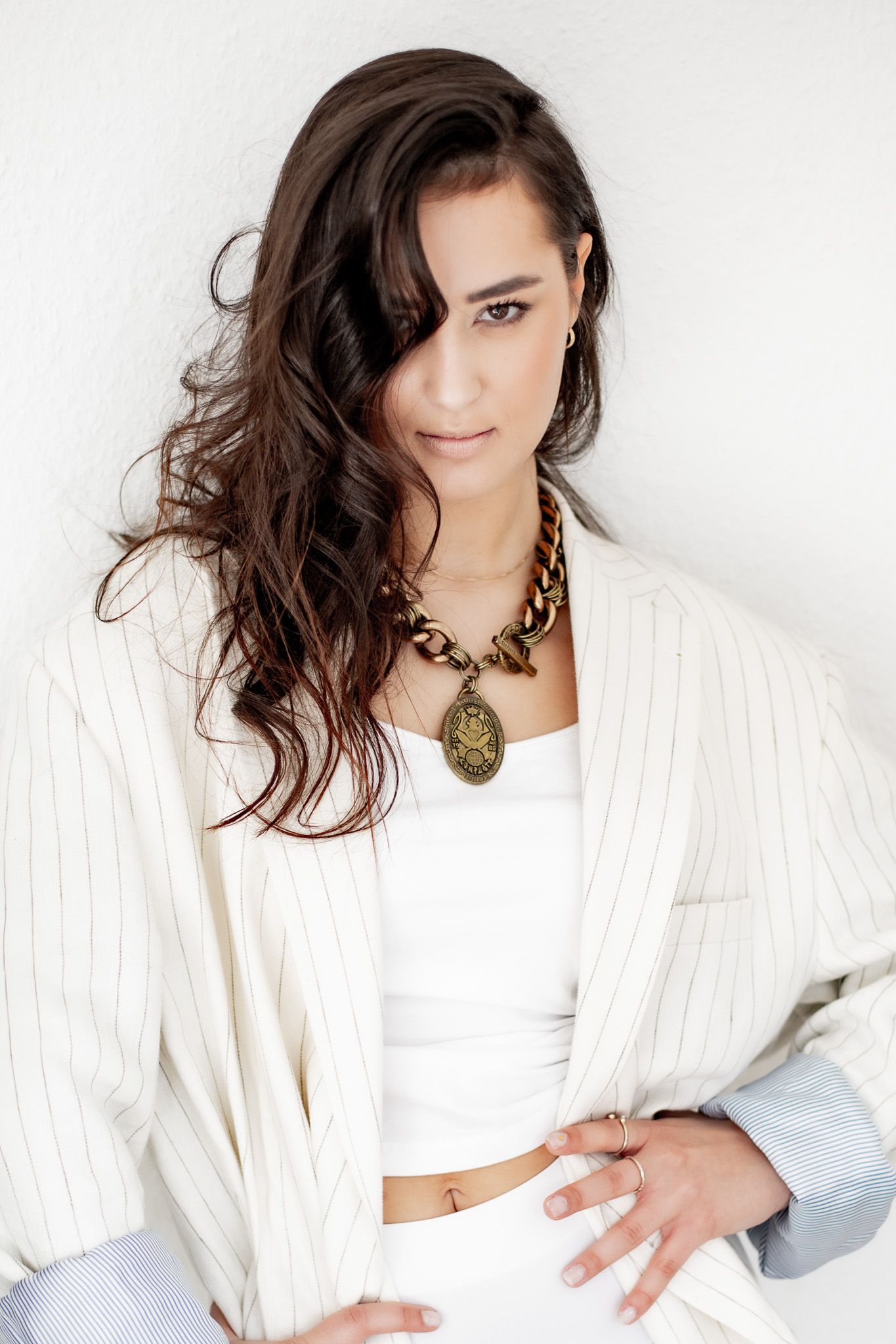 Stylish brunette woman in white striped blazer and gold chain necklace posing against white wall.