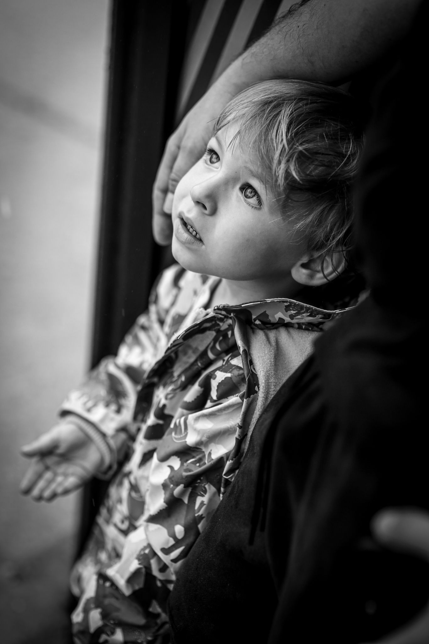 Black and white portrait of a young child looking up with a gentle smile, wearing a camouflage jacket.