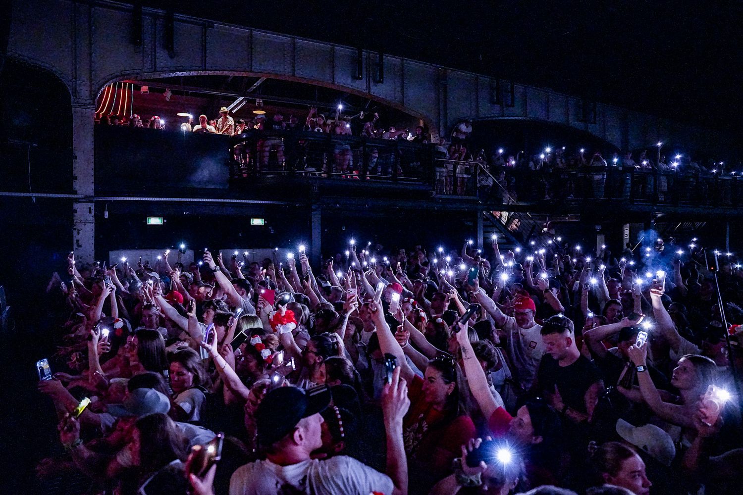 Large concert crowd at night with hands raised holding glowing phone lights under a dark sky.Polonia music festival