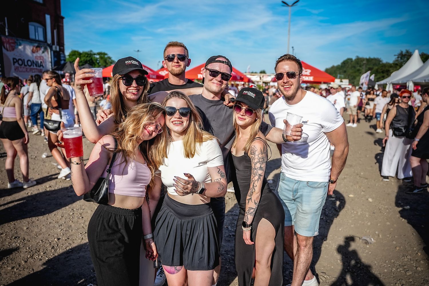 Group of friends posing together at an outdoor summer festival, smiling and holding drinks on a sunny day.