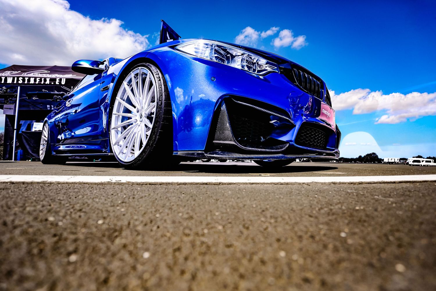 Blue BMW M4 with wide body kit and custom wheels photographed low angle on a racetrack.